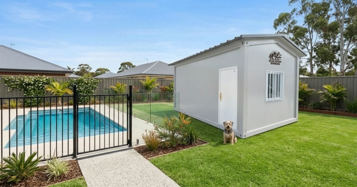 A Dead Dingo Donga insulated garden shed installed on the lawn of a landscaped suburban backyard beside a fenced swimming pool, with a small dog sitting by the door